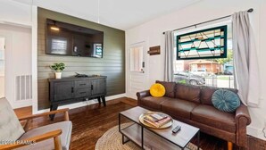 A living room with a dark brown leather sofa, a coffee table with books and a remote, a black TV on a green wall, and a window with curtains.