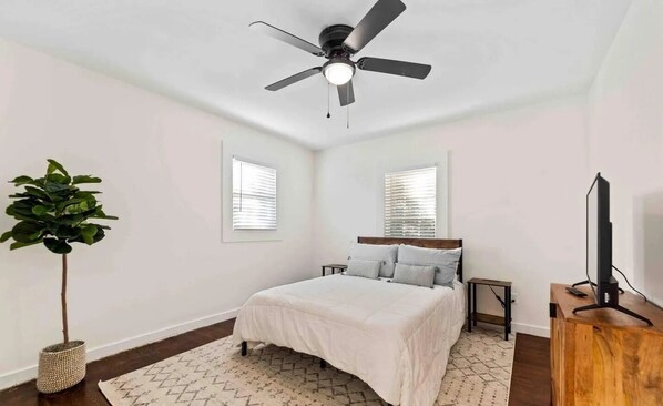 A bedroom with a bed, a ceiling fan, a TV, and a plant.