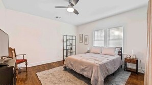 A bedroom with a bed, a nightstand, a desk, a chair, a ceiling fan, and a window with blinds.