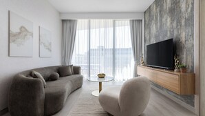 A modern living room with a grey sofa, a round armchair, a flat-screen TV on a wooden console, and large windows with sheer curtains.