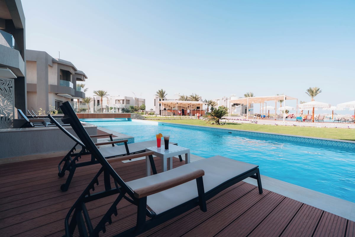 A poolside area with lounge chairs and a table, overlooking a swimming pool and resort buildings.