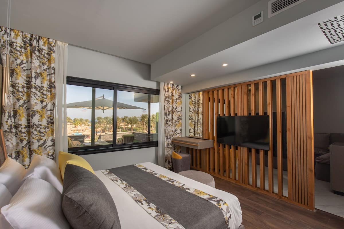 A modern hotel room with a large bed, a TV mounted on a wooden panel, and a view of the outdoors through a large window.