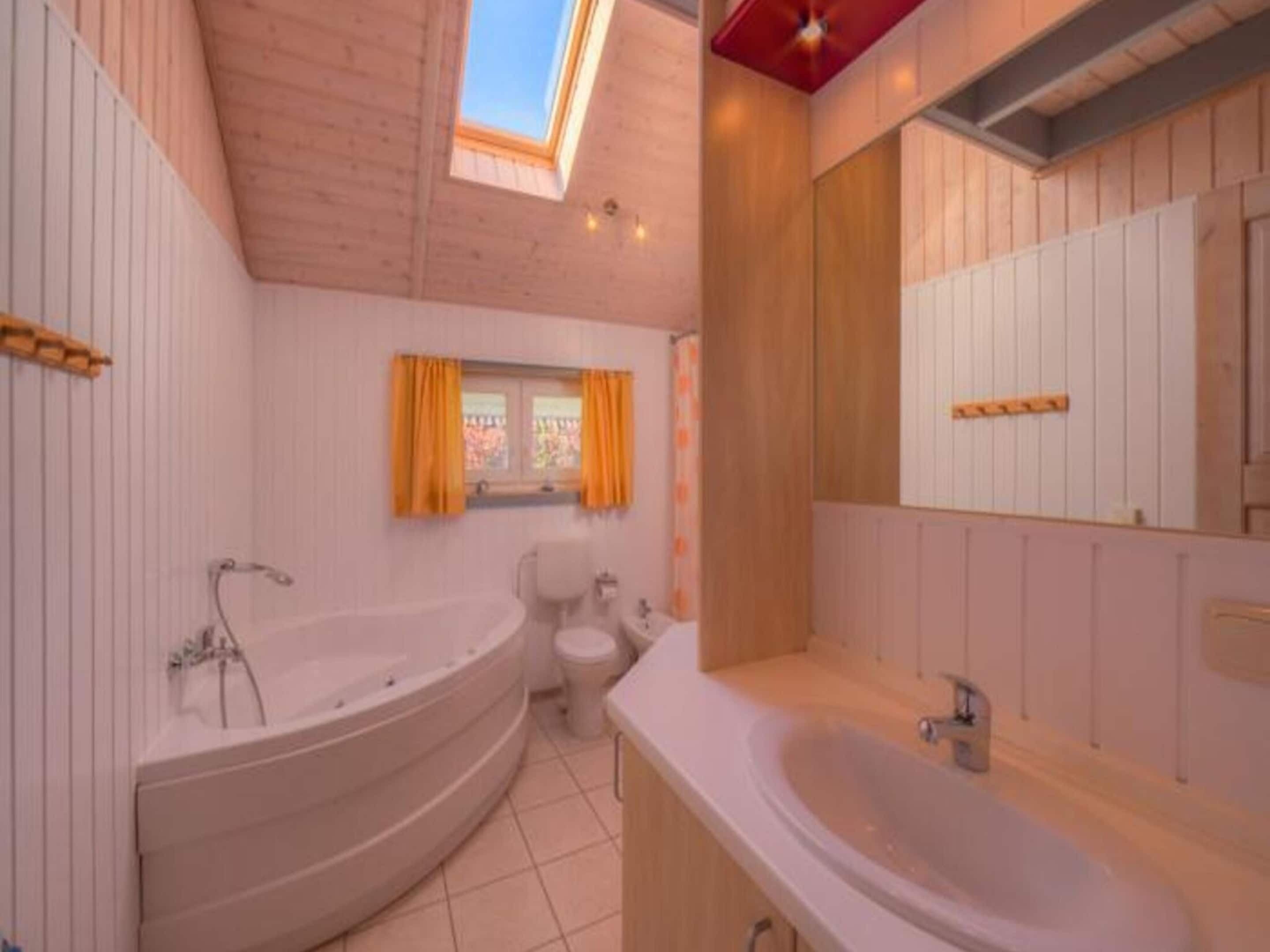 A modern bathroom with a freestanding bathtub, a toilet, and a large sink. There is a skylight above the bathtub and a window with curtains.