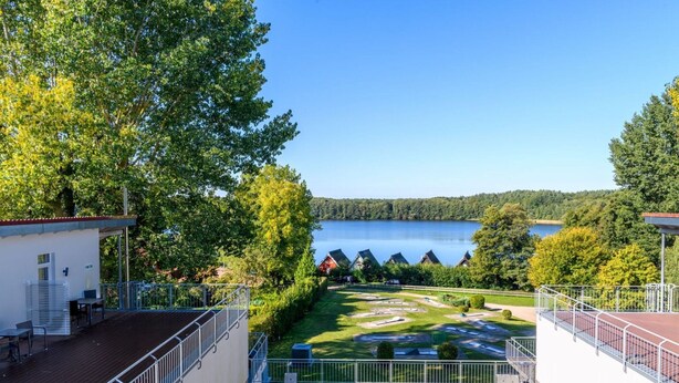 Vista da un balcone su un lago, con una zona verde e un edificio con tetto rosso.