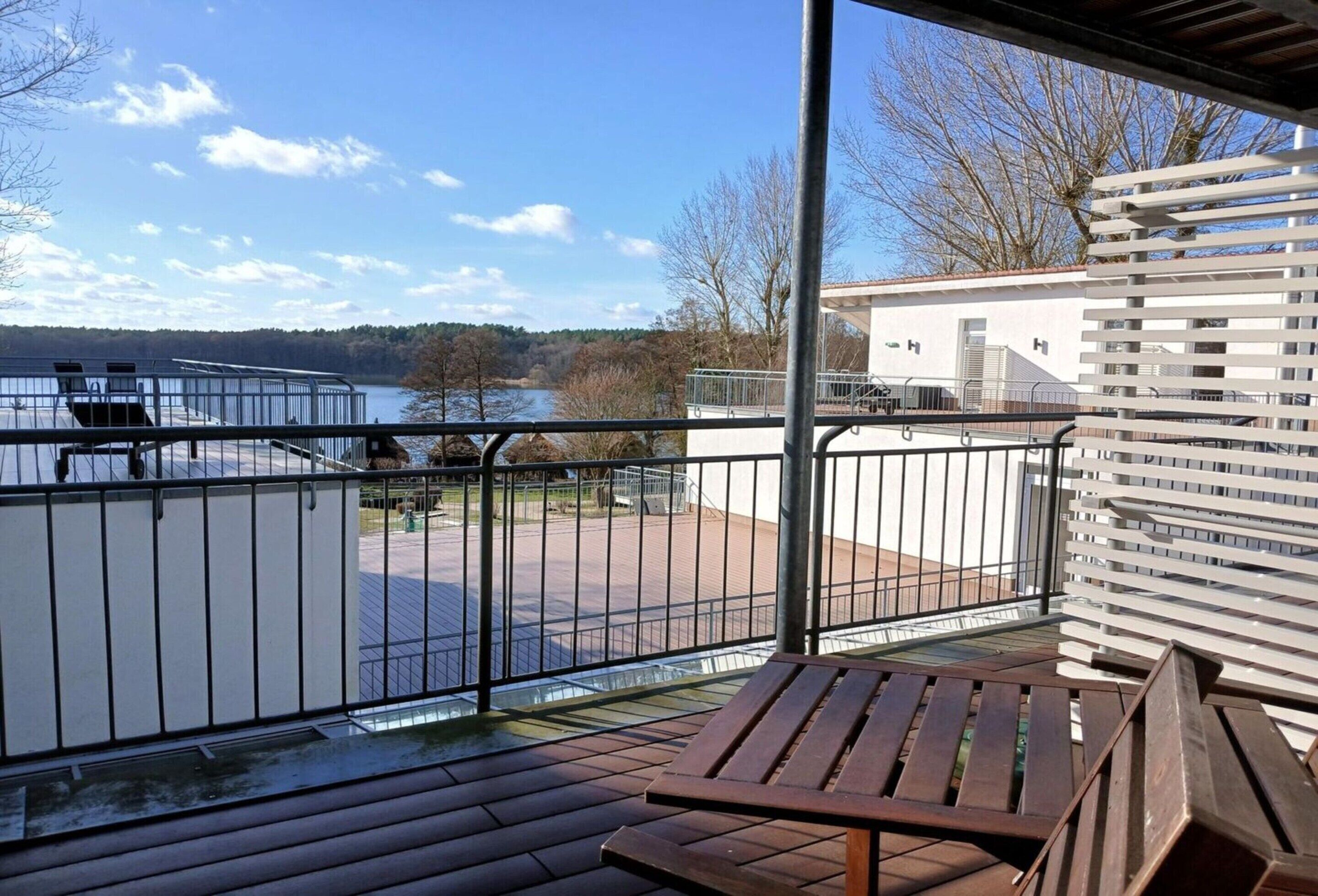 Un balcon donnant sur un lac et des arbres, un banc en bois et, au loin, un bâtiment blanc.
