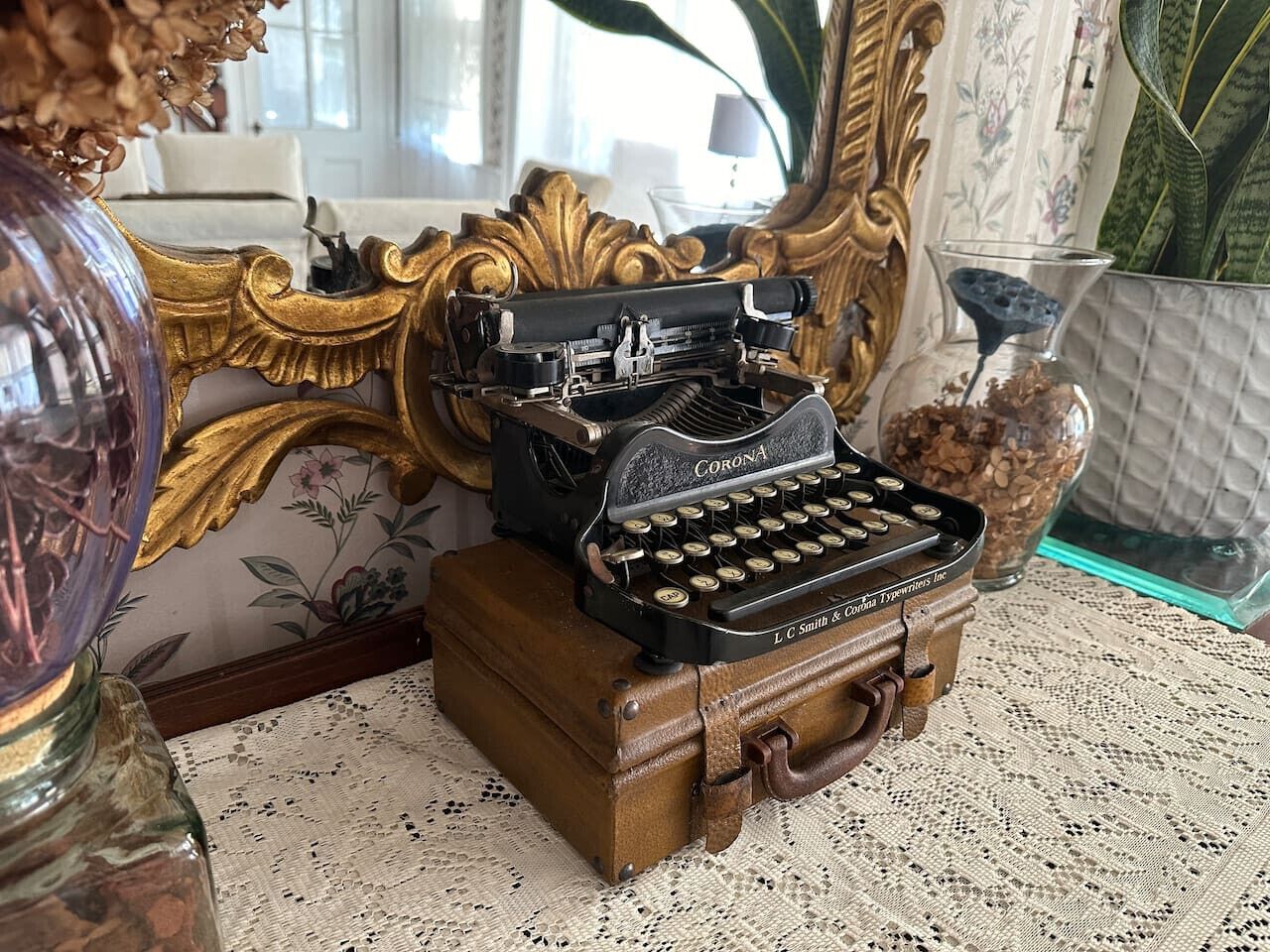 A vintage Corona typewriter on a wooden box, a lace tablecloth, and a mirror with floral wallpaper.