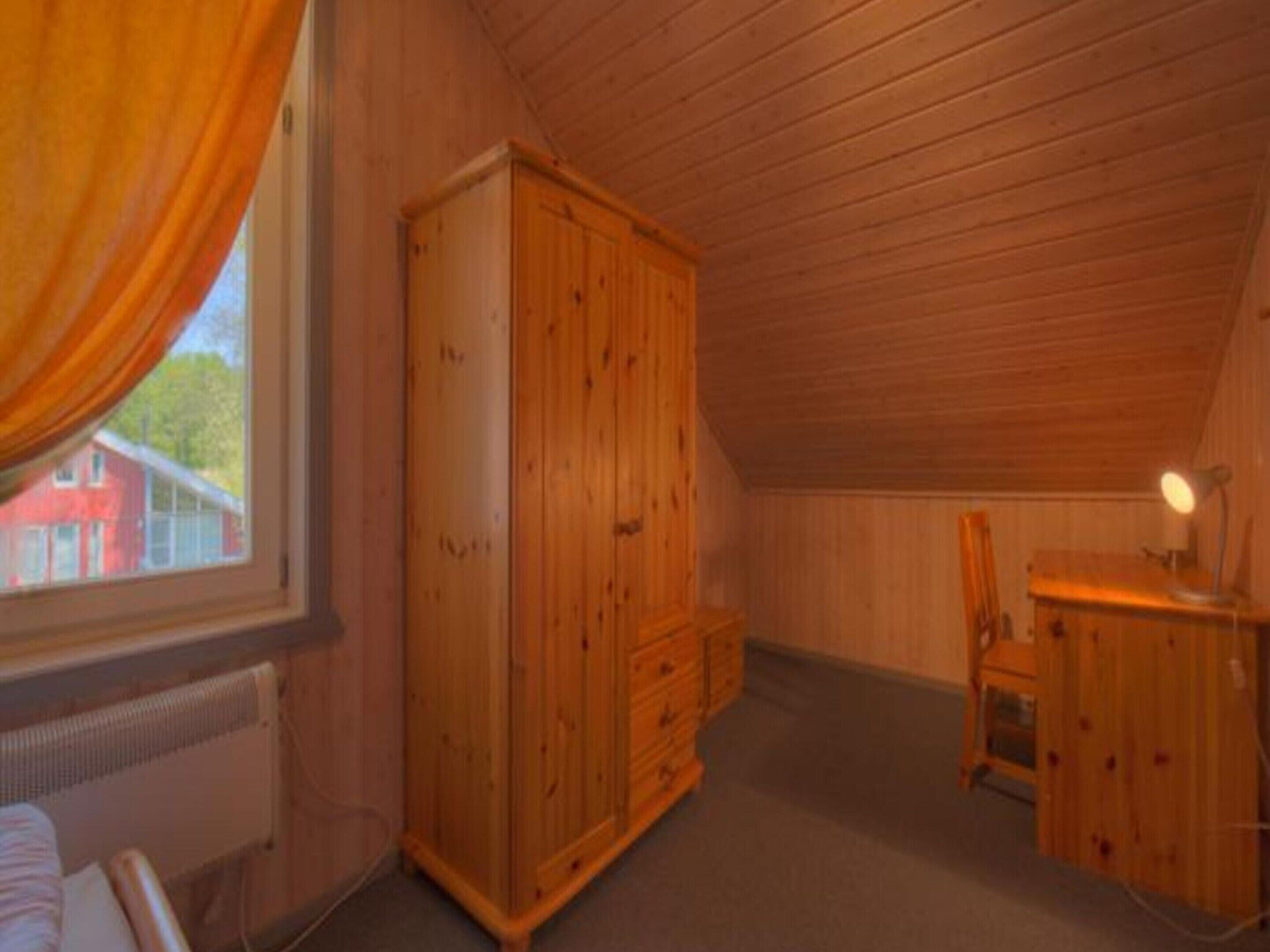 Ein gemütliches Dachzimmer mit Holzwänden und schräger Decke. Es beinhaltet einen großen Holzschrank, einen Schreibtisch mit Lampe sowie einen Stuhl.