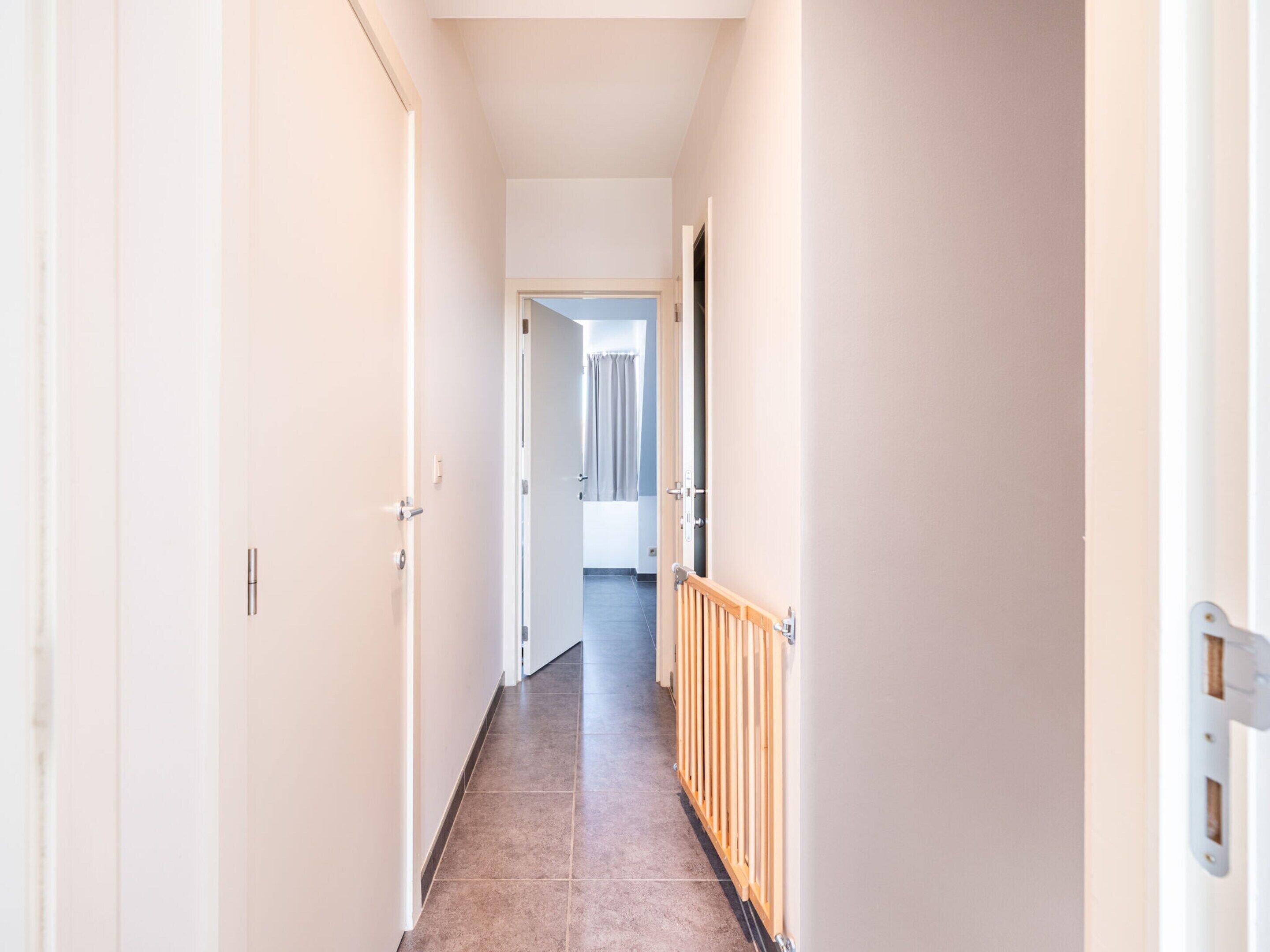 Un couloir étroit aux murs blancs et au sol carrelé, qui mène à une pièce dotée d’une porte et d’une fenêtre.