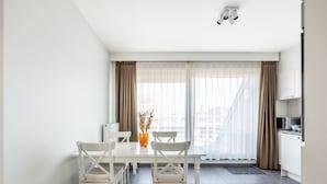 Una habitación moderna y luminosa con una mesa de comedor y sillas, un radiador, una ventana con cortinas translúcidas y una kitchenette.