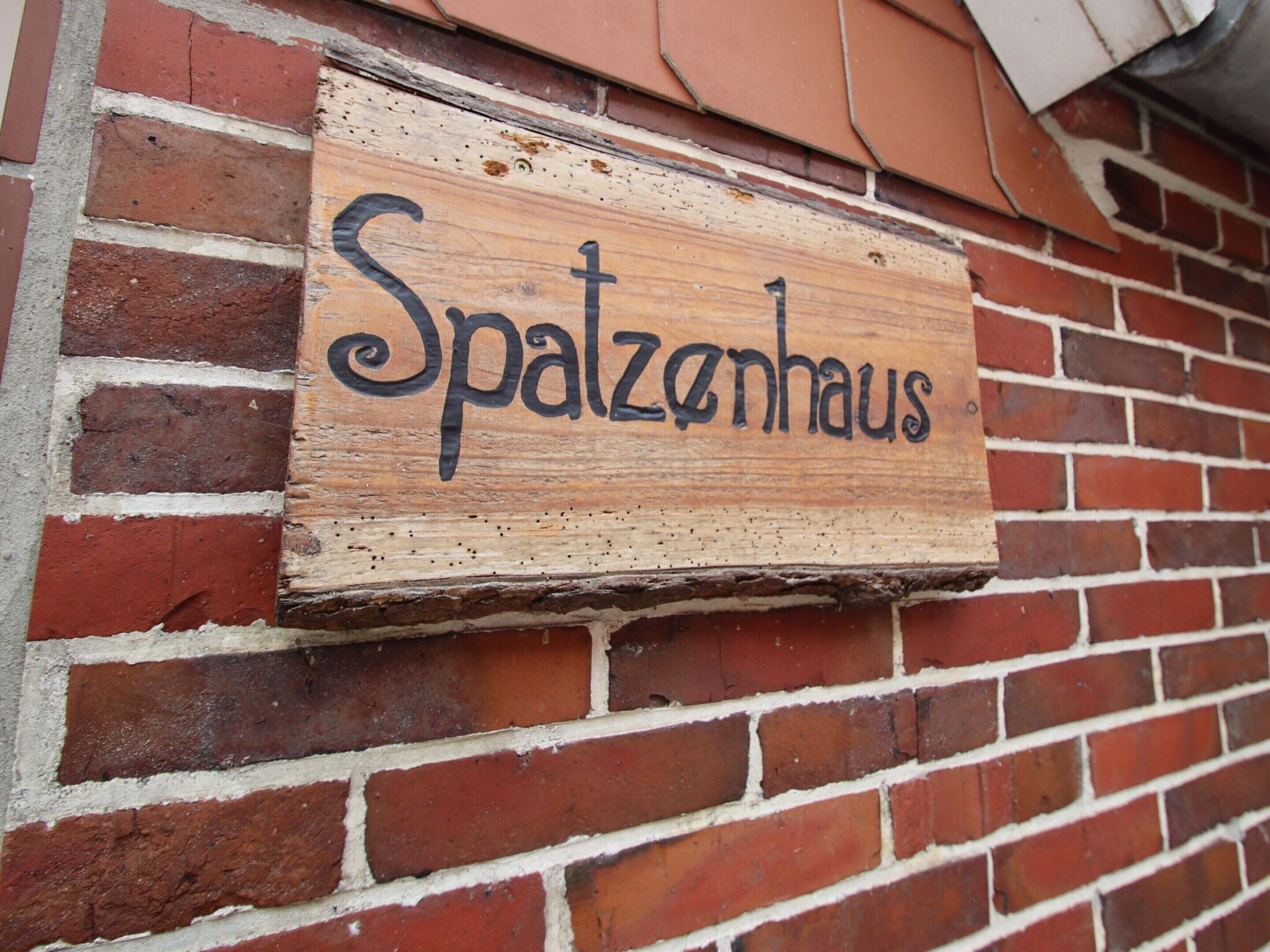Une enseigne en bois portant l’inscription « Spatzenhaus » est fixée à un mur de briques.