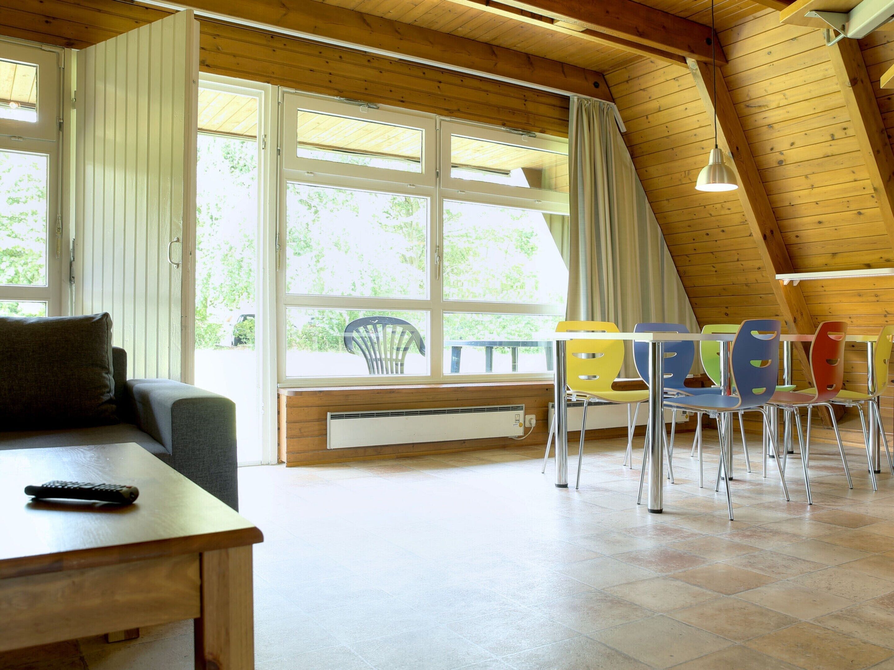 Une pièce chaleureuse avec un plafond en bois, un canapé, une table, des chaises colorées et de grandes fenêtres.