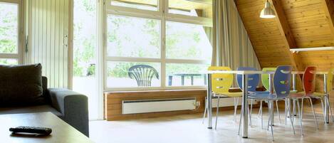 Une pièce chaleureuse avec un plafond en bois, un canapé, une table, des chaises colorées et de grandes fenêtres.