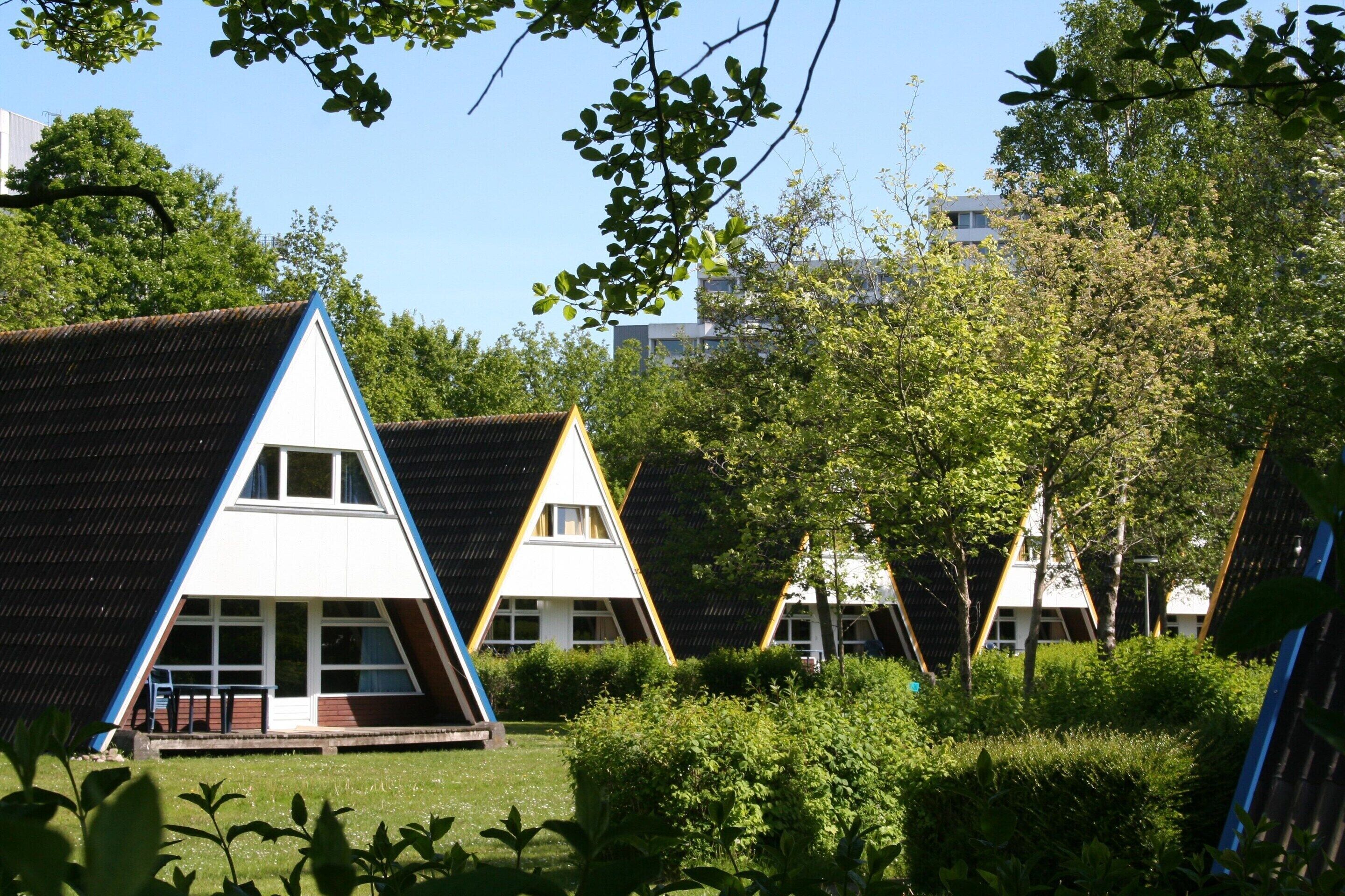 Een rij vakantiehuisjes met puntige daken, een gazon ervoor, omringd door bomen en een heldere lucht.