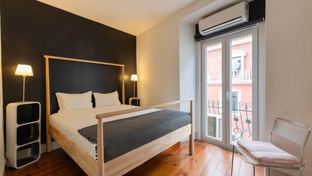 Un dormitorio con cama, lámparas de noche, equipo de aire acondicionado, ventana con vista a un balcón y una silla.