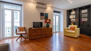 Amplia sala de estar con suelo de madera, un sofá, una silla, un televisor sobre un mueble de madera y un balcón con barandilla.