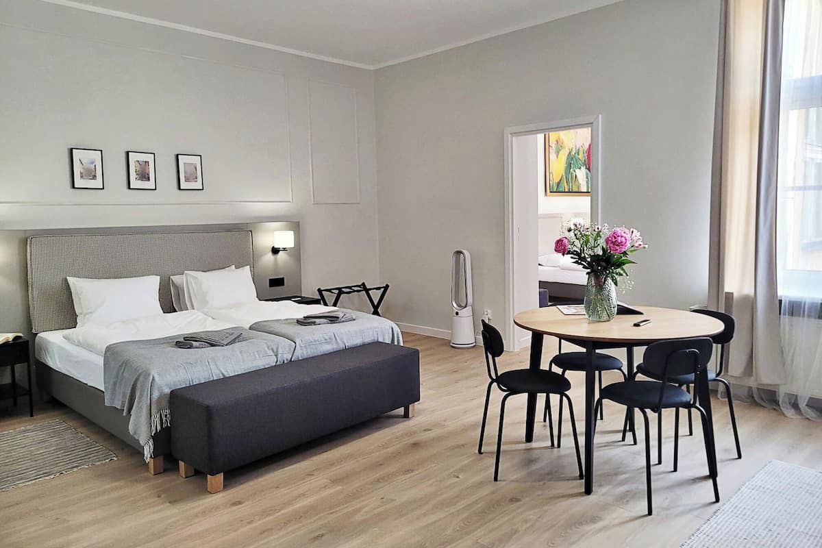 Quarto de hotel moderno com cama, mesa de cabeceira, luminária, espelho sobre a mesa e um espaço de refeições com cadeiras e uma mesa.