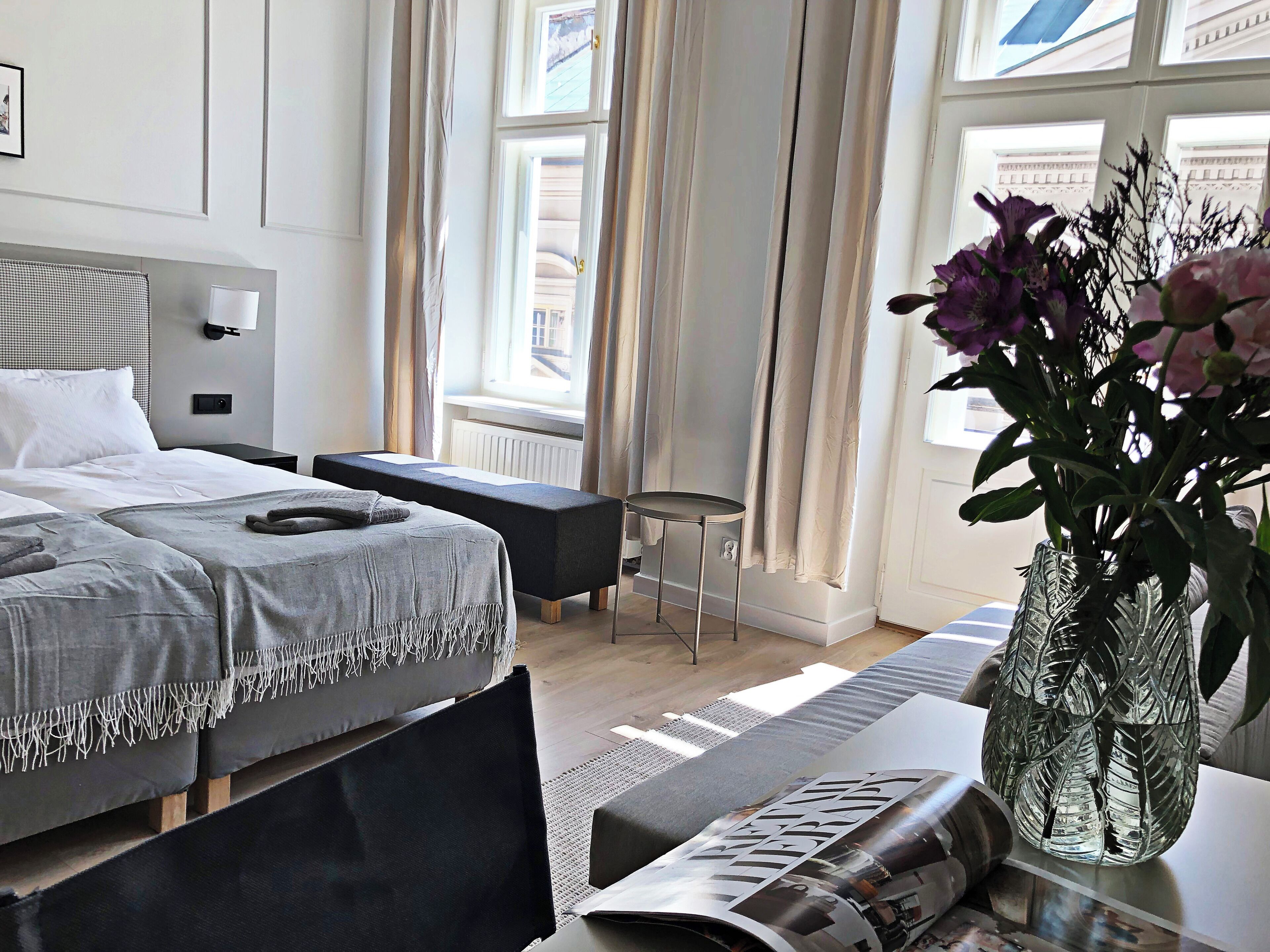 A modern hotel room with a neatly made bed, a small table with a vase of flowers, and a window with curtains.