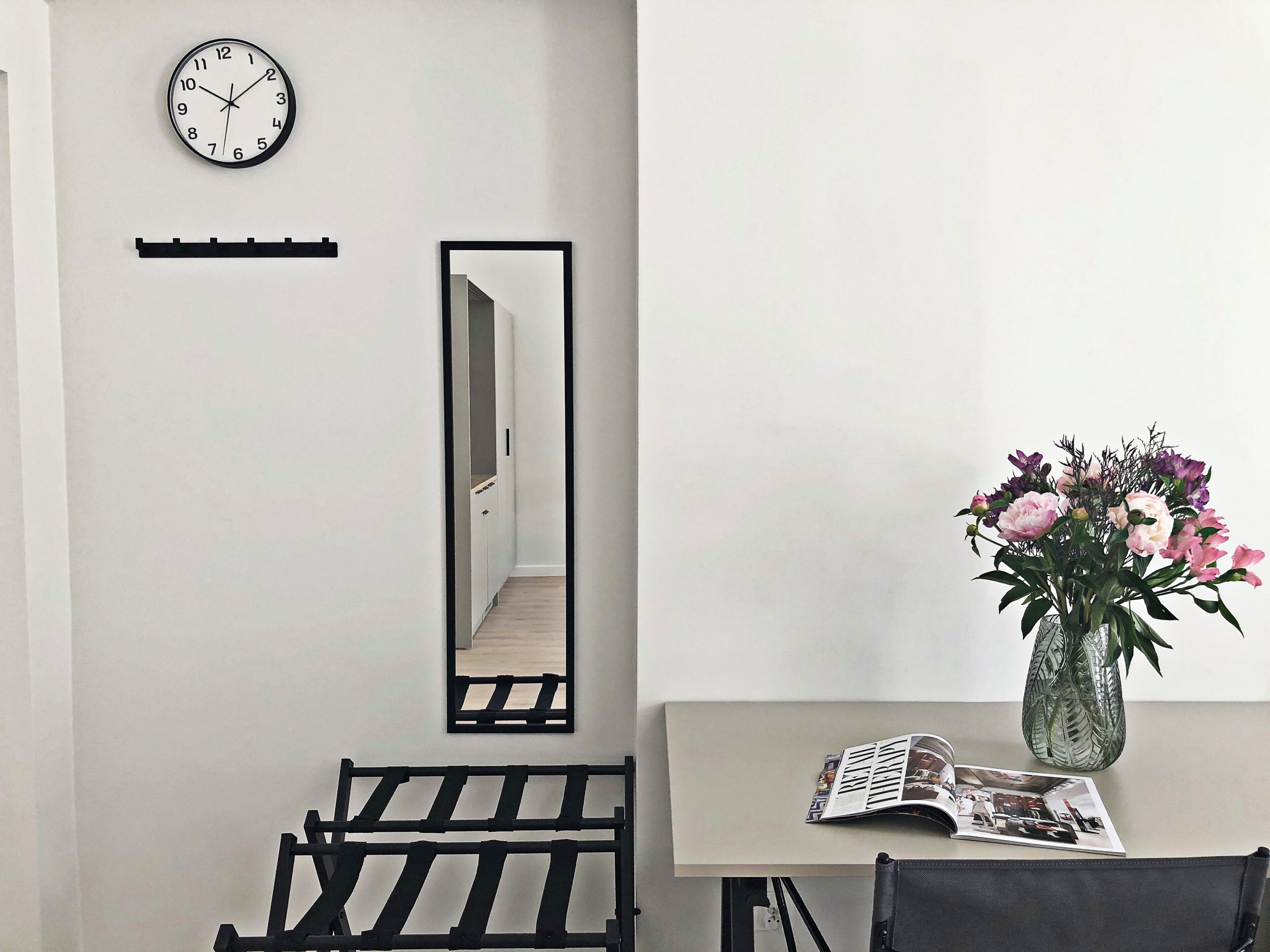 Een kamer met een wandklok, een spiegel, een bankje, een tafel met een bloevas en een tijdschrift.