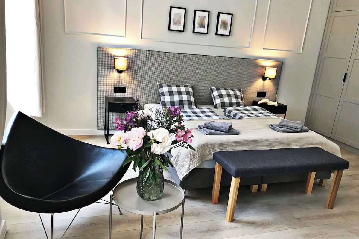 Um quarto moderno com uma cama grande, duas mesas de cabeceira, uma cadeira, uma mesinha com um vaso de flores e um banquinho.