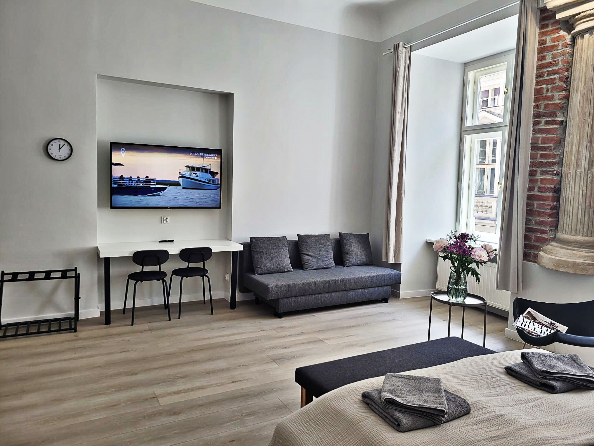 Moderni hotellihuone, jossa on harmaa sohva, pieni pöytä ja kaksi tuolia, seinälle asennettu televisio ja ikkuna, josta on näkymä ulos.