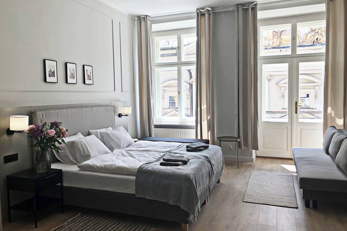 Quarto de hotel com uma cama grande, duas mesas de cabeceira, um criado-mudo, um sofá e vista da cidade através de amplas janelas.
