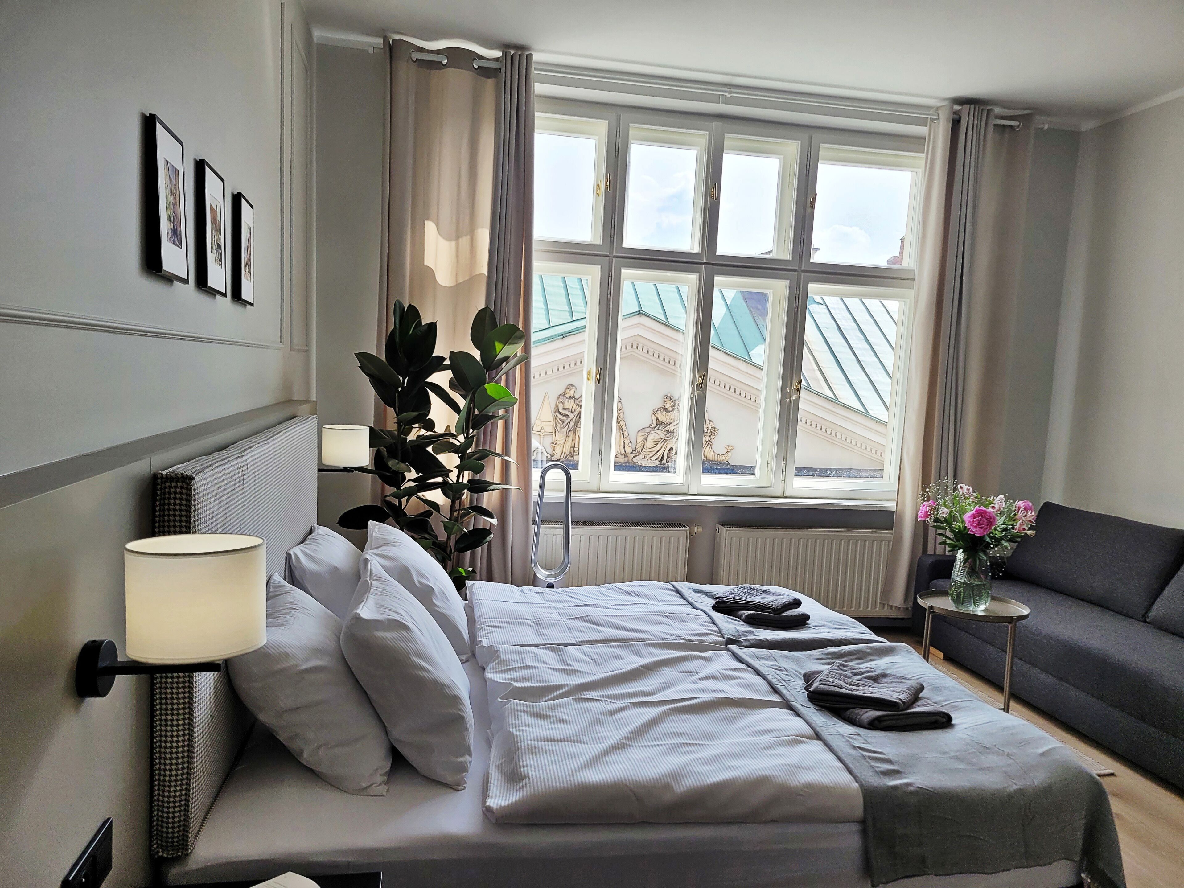 Habitación de hotel con cama, lámpara de noche, planta en maceta, ventana con vista a un edificio y un sofá.