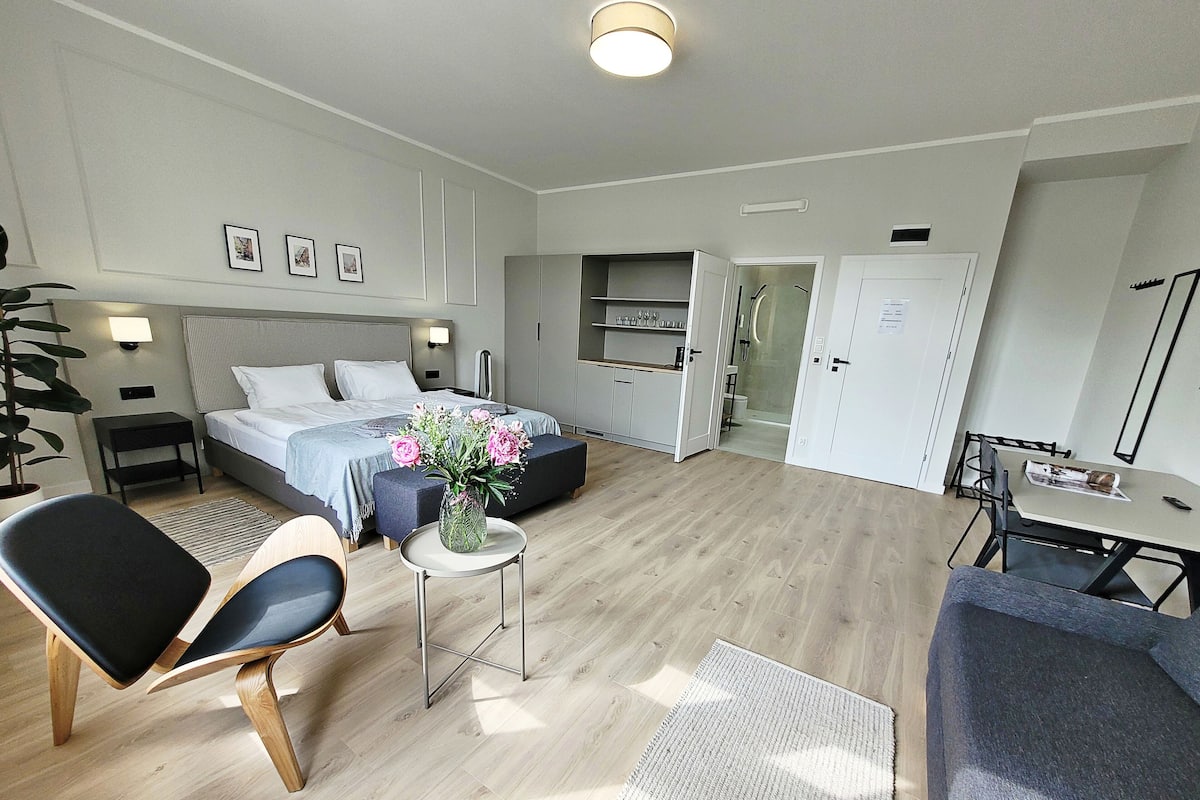 Quarto de hotel moderno com uma cama, mesas de cabeceira, uma escrivaninha, uma cadeira, uma pequena mesa com um vaso de flores e área de chuveiro.