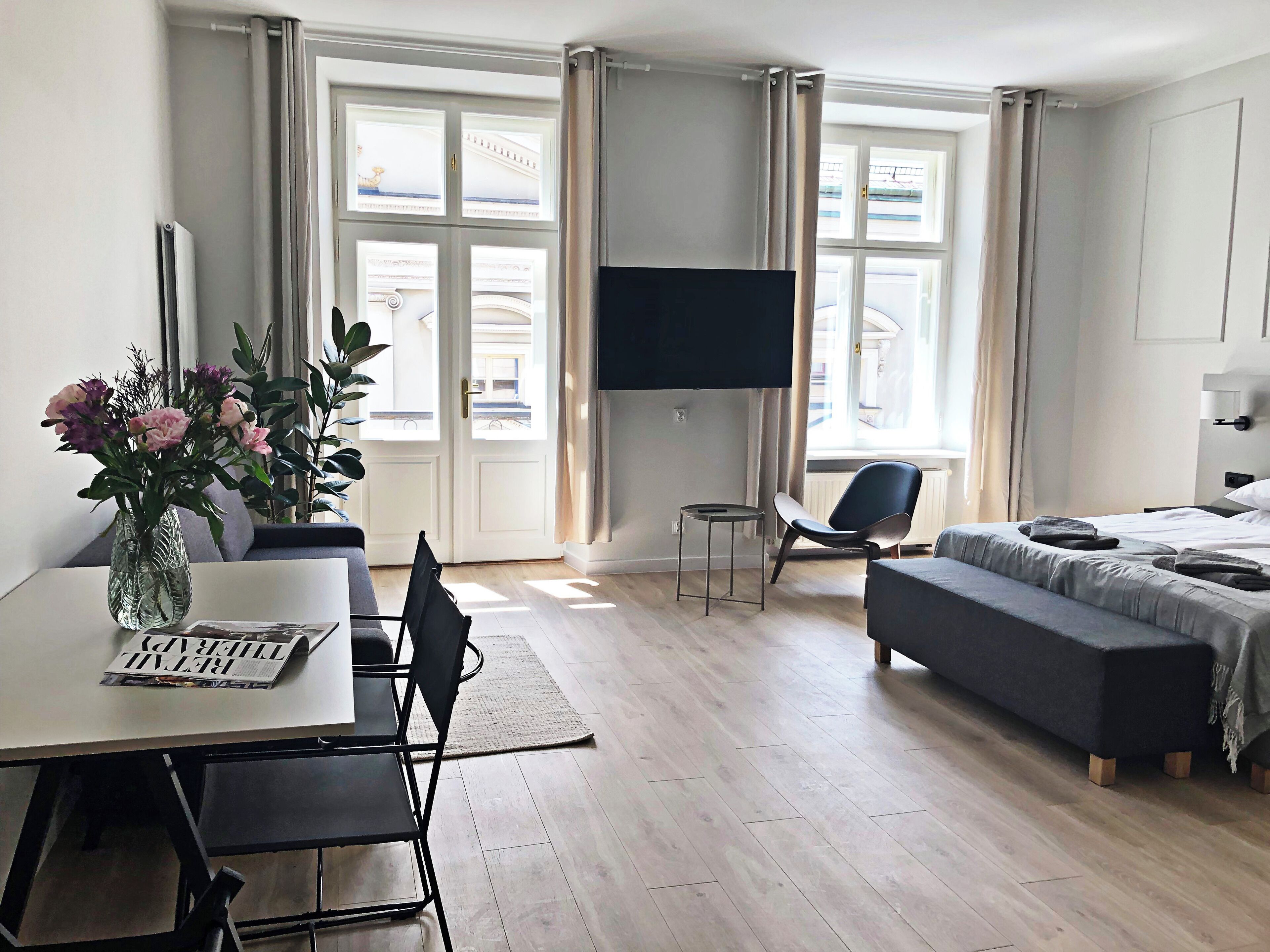 Una habitación de hotel moderna con zona de comedor, sofá, televisión y ventana con cortinas.