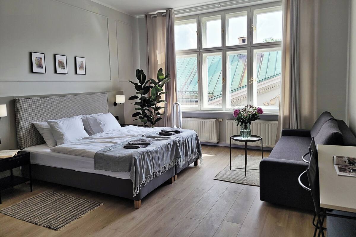 Quarto de hotel moderno com uma cama grande, um pequeno sofá, uma escrivaninha e uma janela com vista para a paisagem urbana.