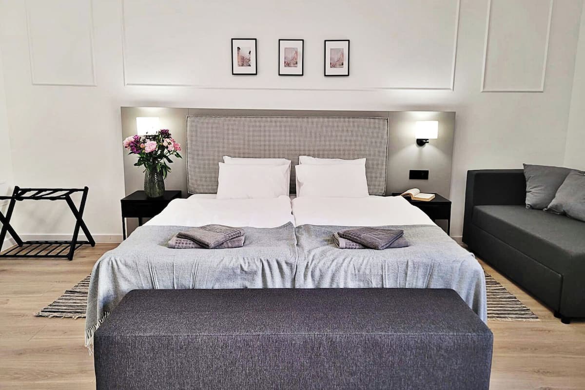 Quarto de hotel moderno com uma cama grande, uma mesa de cabeceira com abajur, uma pequena mesa com um vaso de flores, um sofá e um banquinho.