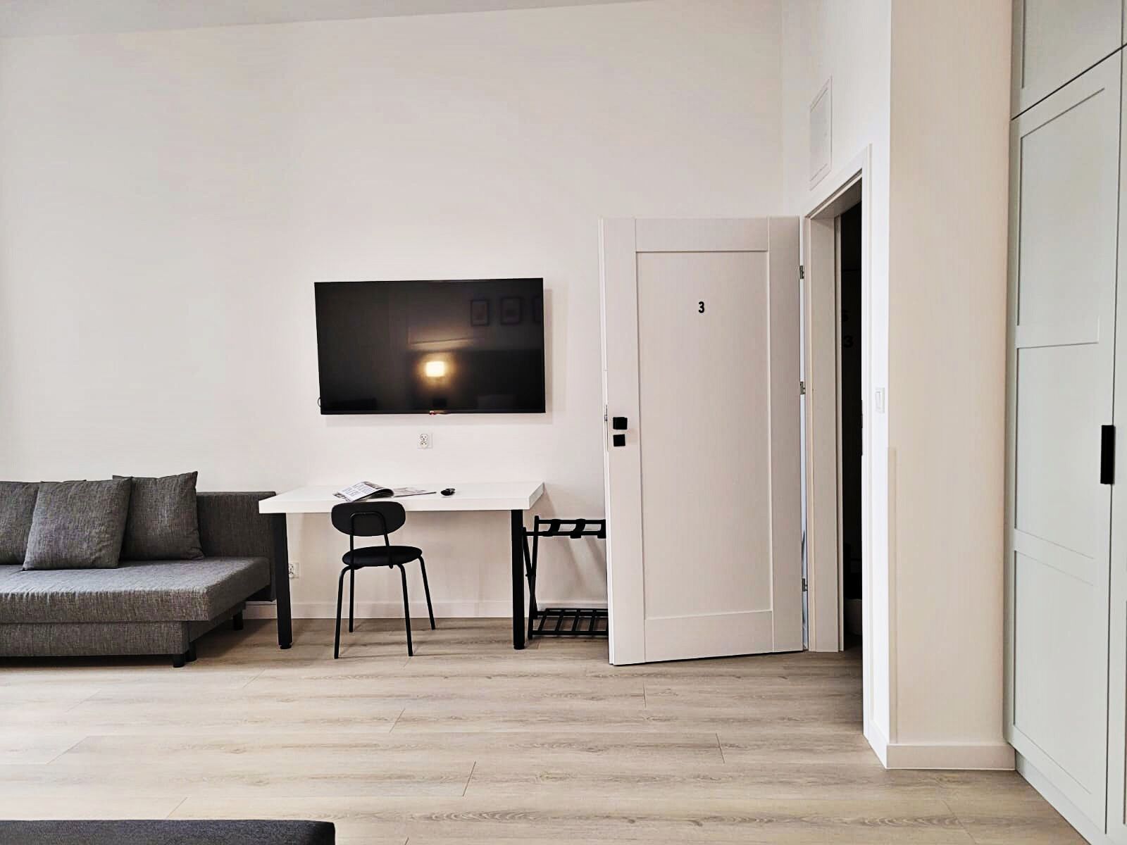 Een moderne woonkamer met een grijze bank, een wit bureau, een zwarte stoel en een flatscreen televisie aan de muur.