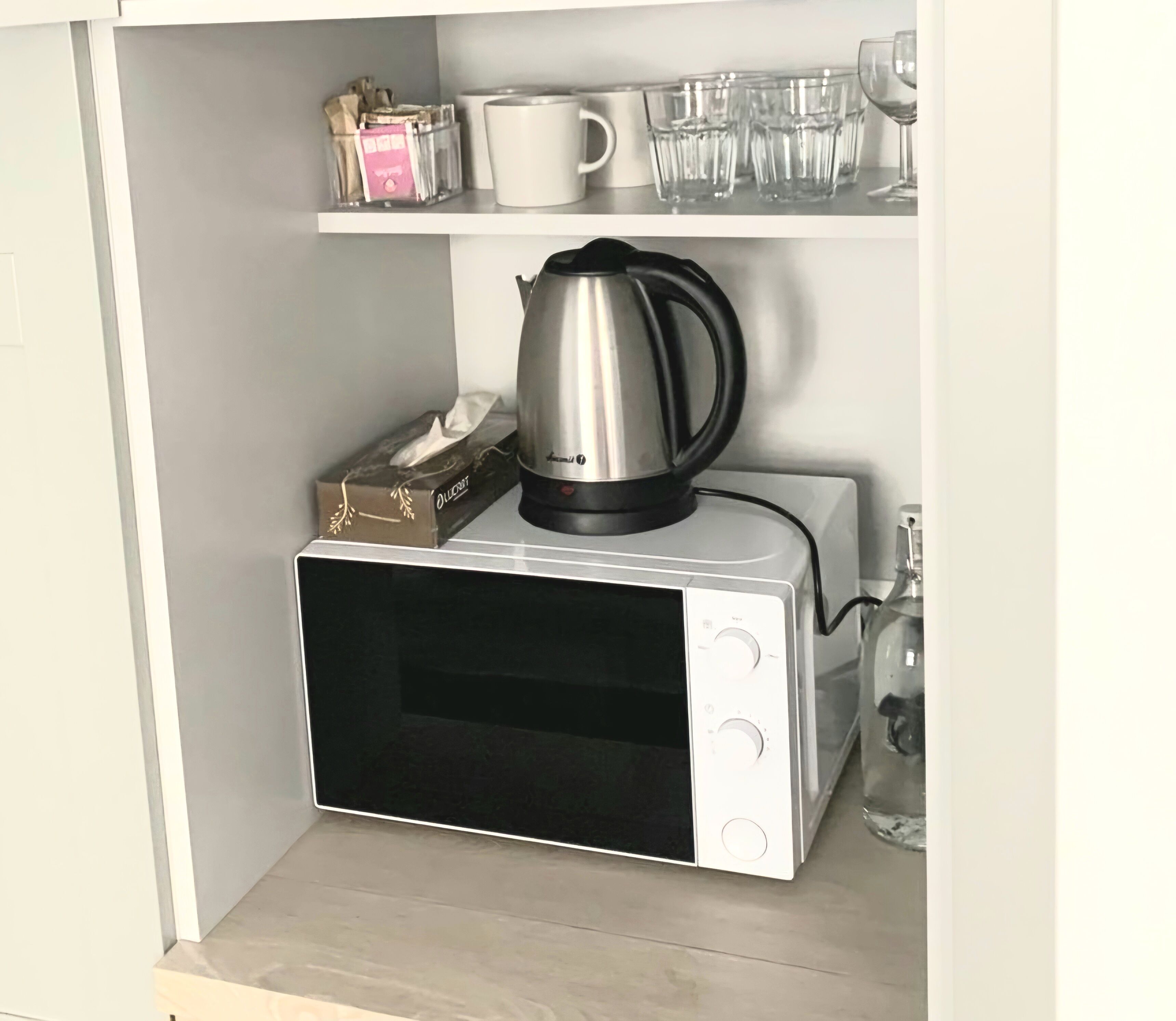 A compact microwave oven with a kettle on top, a tissue box, and a glass set on a shelf above.