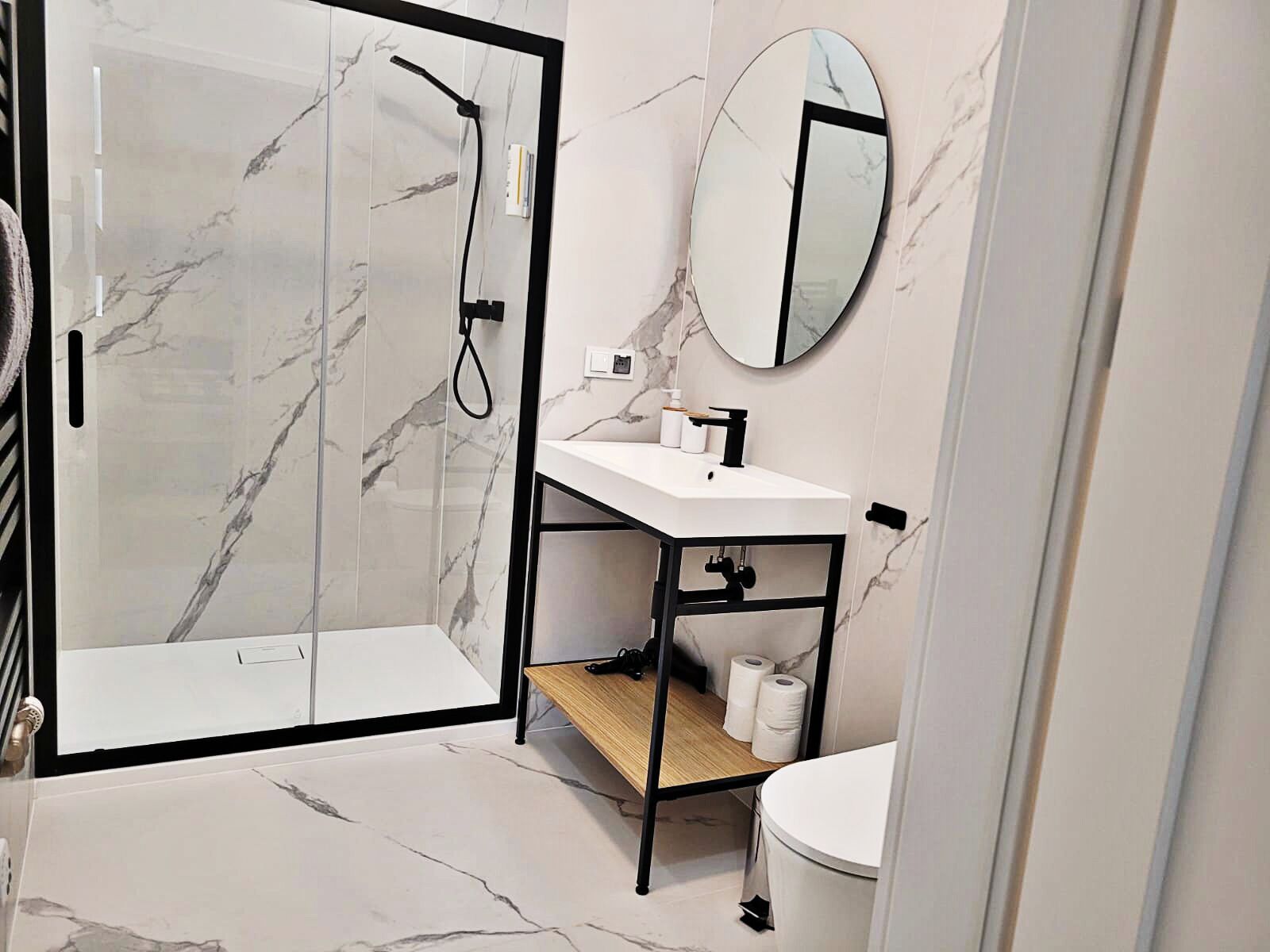 Un baño moderno con ducha envuelta en vidrio, un lavamanos blanco con grifo negro y un espejo redondo en la pared.