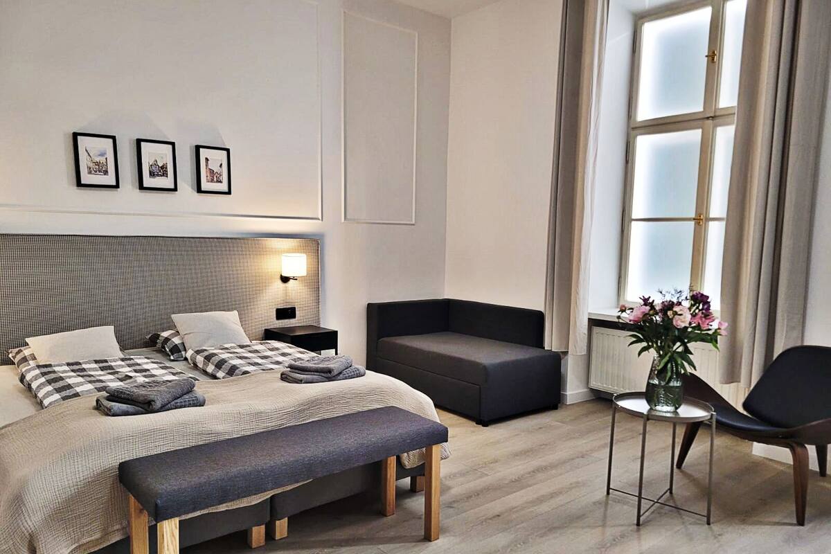 Quarto de hotel moderno com uma cama grande, um banco, um sofá, uma mesinha com um vaso de flores e uma janela com cortinas.