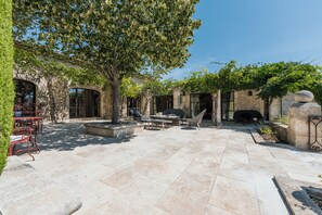 Un patio con suelo de piedra, mobiliario exterior, una chimenea y una pérgola con plantas trepadoras.