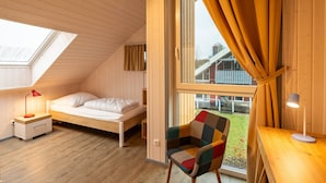 A small, cozy room with a sloped ceiling, a single bed, a chair, a desk, and a large window with curtains. - Ferienhaus im Skandinavischen Stil (Mirow)