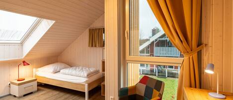 Een kleine, knusse kamer met schuine wanden, een enkel bed, een stoel, een bureau en een groot raam met gordijnen.