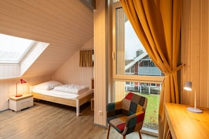 A small, cozy room with a sloped ceiling, a single bed, a chair, a desk, and a large window with curtains. - Ferienhaus im Skandinavischen Stil (Mirow)