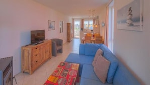 A living room with a blue sofa, wooden cabinet, and a television. There is a dining area in the background with a table and chairs. - Appartement im Ferienpark in Rechlin (Rechlin)