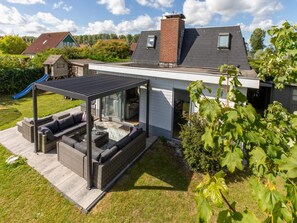 Ein modernes Haus mit überdachter Terrasse, einem sichtbaren Schornstein und einem Garten mit einer Rutsche.