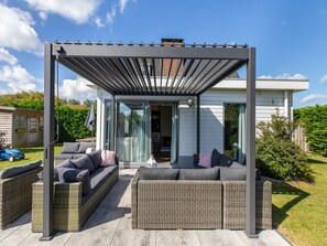 Ein überdachter Außenbereich mit einer Couch, Stühlen, einer Pergola und einer Glastür zum Haus.