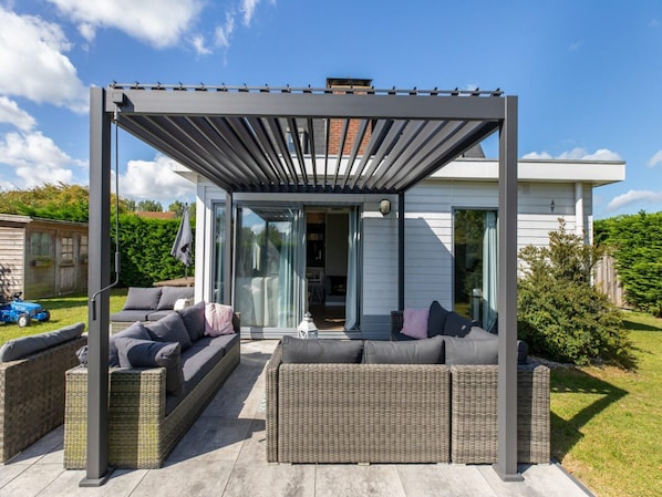 A covered outdoor seating area with a sofa and wicker furniture, a pergola, and a house in the background.