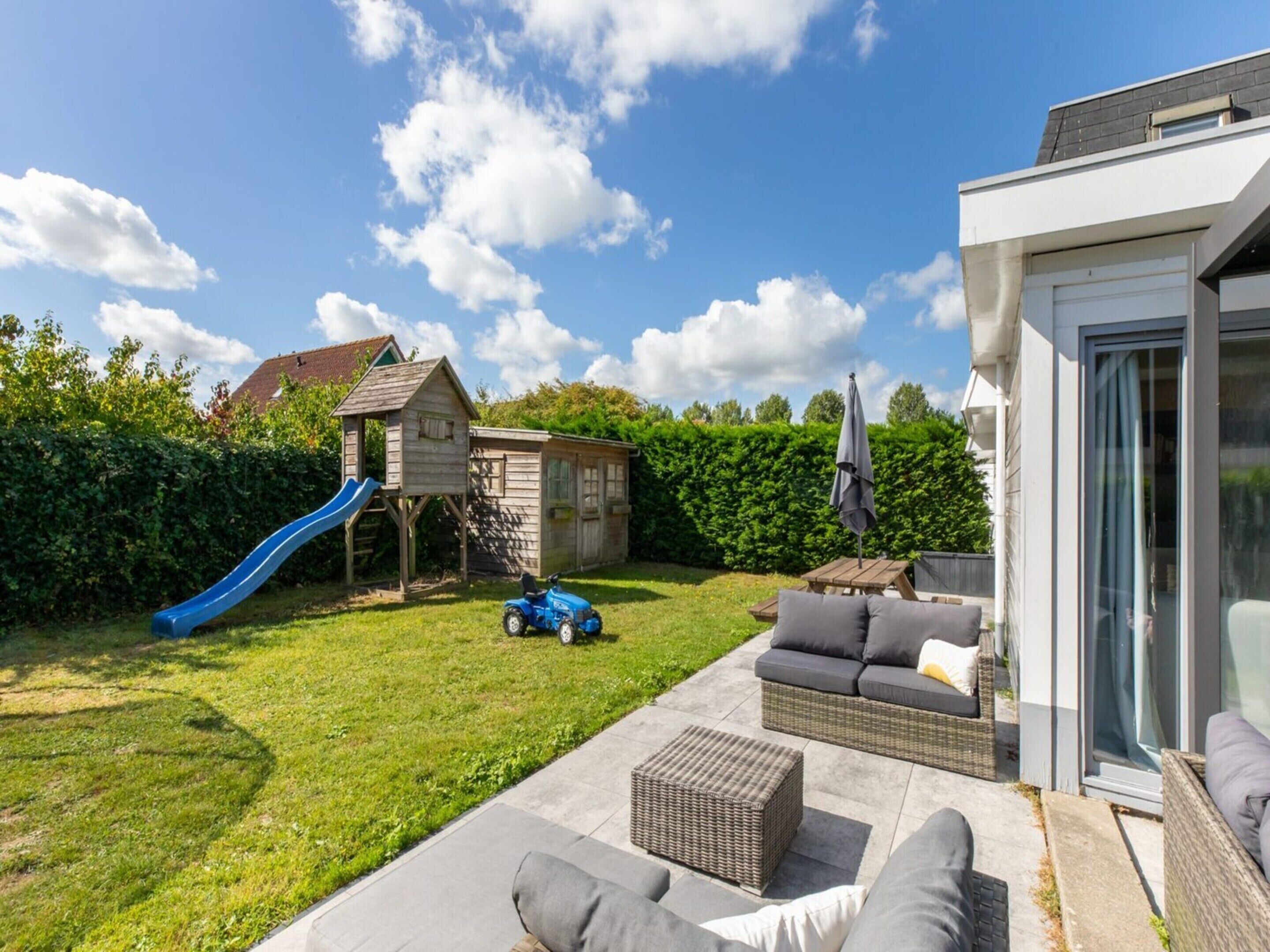 A backyard with a playhouse, a slide, a blue toy car, and outdoor furniture.