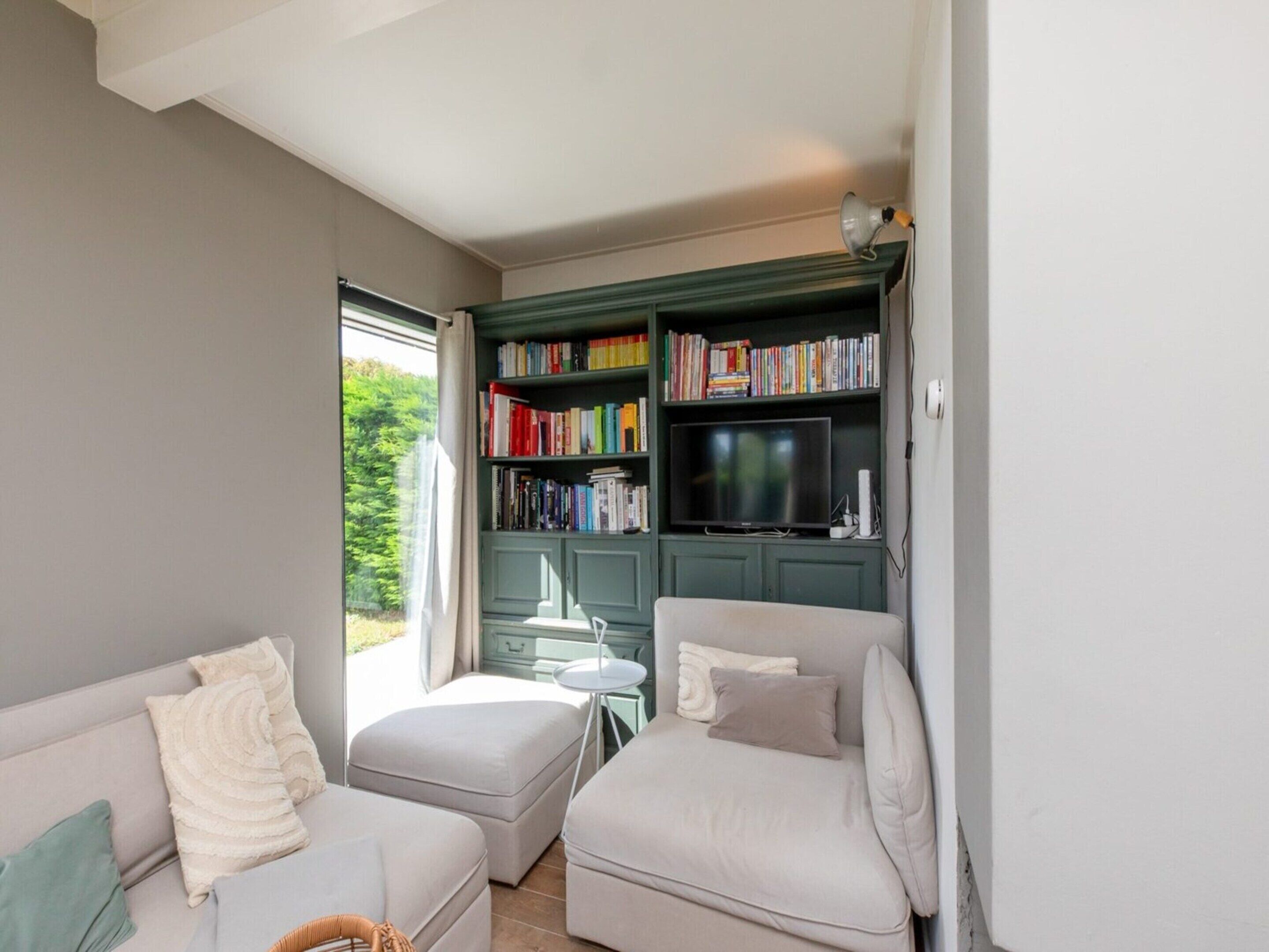 A living room with a sofa, ottoman, bookshelf, television, and a view of greenery outside.