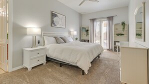 Un dormitorio con cama, mesitas de noche, cómoda, ventilador de techo, ventana con cortinas y estante en la pared con plantas.