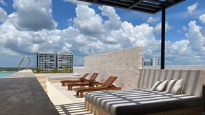 Un dormitorio moderno al aire libre con cama, sillones de descanso y vistas al mar y edificios.
