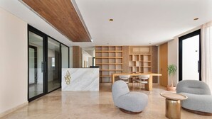 Un vestíbulo moderno con un mostrador de recepción de mármol, una estantería de madera, un área de comedor con mesa y sillas, y dos sillones grises.