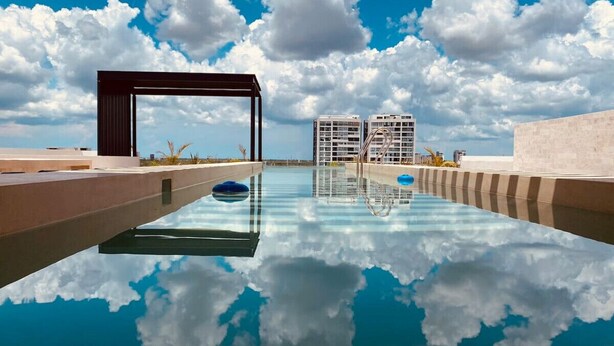 Una piscina con el reflejo nítido de nubes, rodeada de edificios modernos y un cielo despejado.
