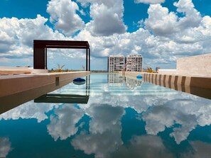A swimming pool with a clear reflection of clouds, surrounded by modern buildings and a clear sky. - Aposento - Private pool, gym & panoramic rooftop by LAHOS (Mérida)