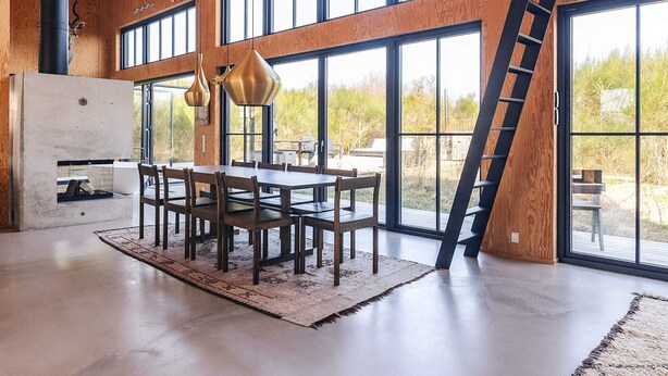 A spacious dining area with a large table and chairs, a concrete fireplace, and large windows offering views of the outdoors. - Chic Retreat in Sjaellands Odde - By Traum Ferienwohnungen (Sjællands Odde)